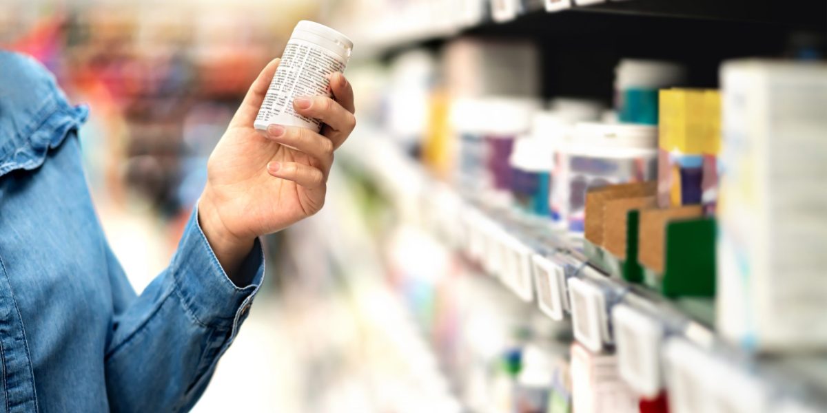 Customer in pharmacy holding medicine bottle. Woman reading the label text about medical information or side effects in drug store. Patient shopping pills for migraine or flu. Vitamin or zinc tablets.