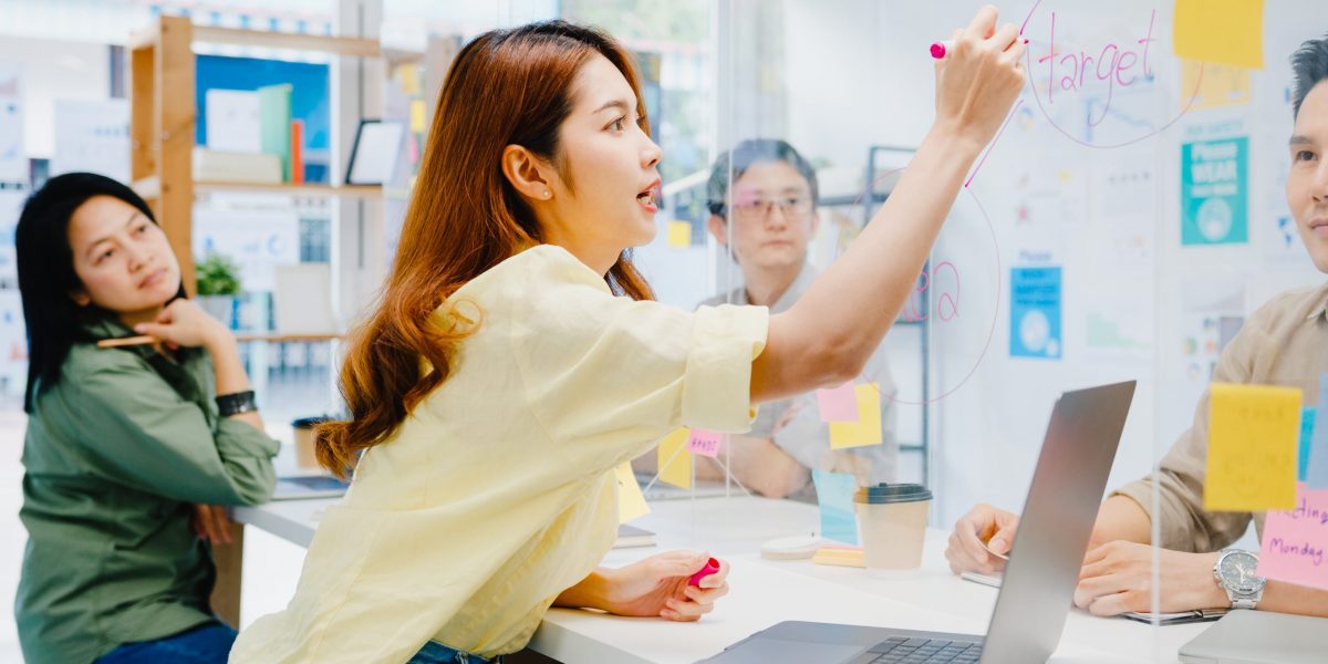Asia businesspeople discussing business brainstorm meeting together share data and write on acrylic partition stand back in new normal office. Lifestyle social distancing and work after coronavirus.