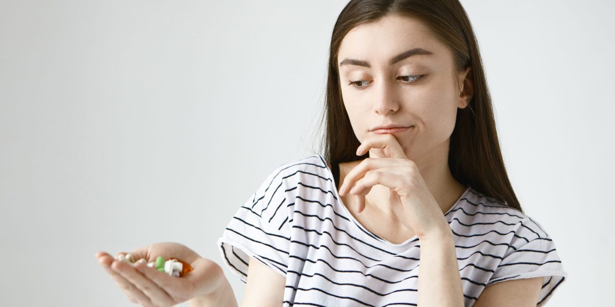 Health, care and treatment concept. Isolated studio shot of indecisive doubtful young lady with loose dark hair rubbing chin thoughtfully and looking at colorful pills on her palm, going to take them